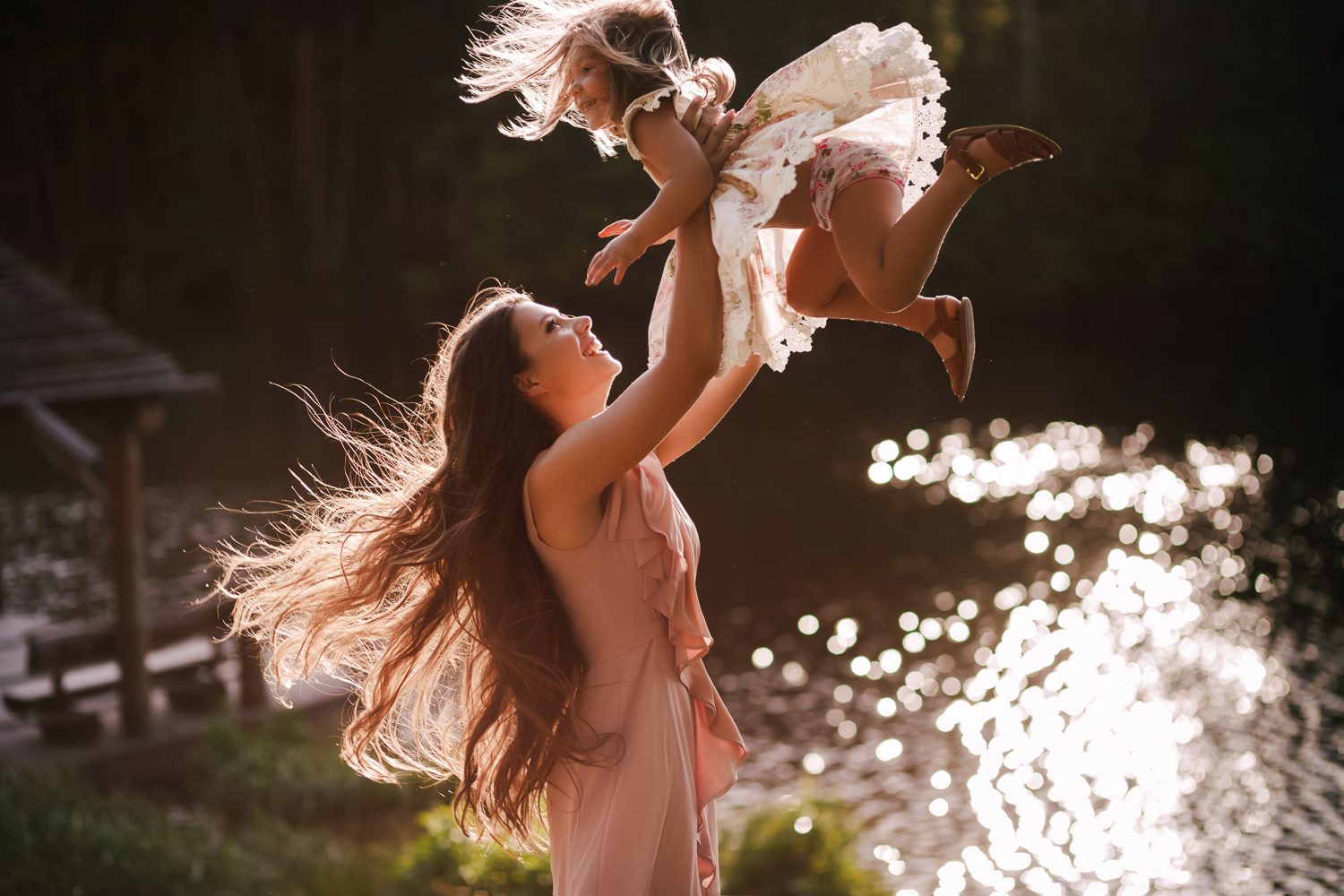 A woman with long flowing hair in a pink dress joyfully lifts a young girl wearing a white dress and brown shoes into the air. They are outdoors near a reflective body of water with sunlight shimmering on its surface. Both appear happy and carefree, perfectly captured by a Charleston family photographer.