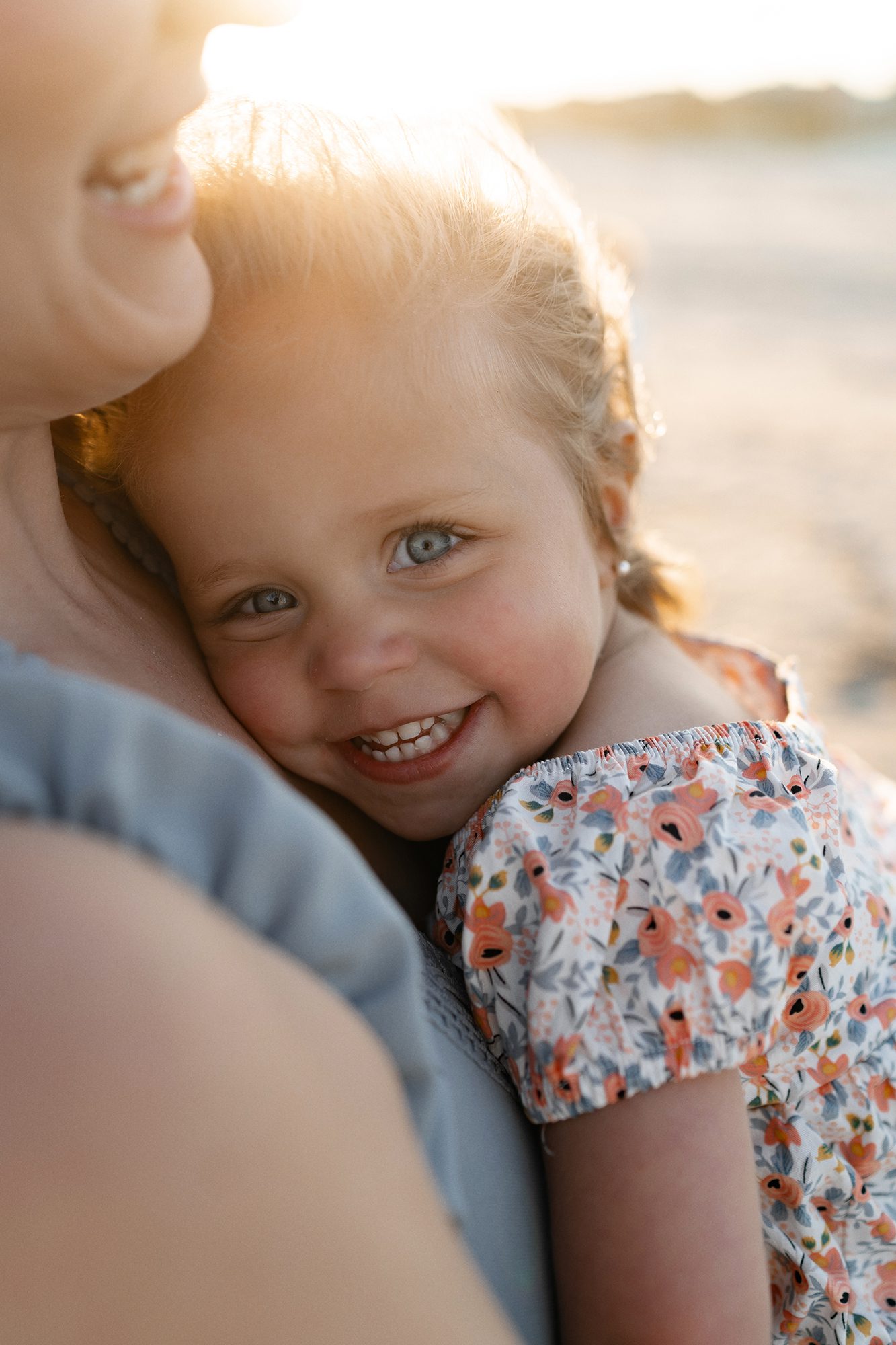 A young child with blue eyes and blonde hair smiles brightly while being held close by an adult. The child is wearing a floral dress with orange and pink flowers. Captured by Olga Chagarov, Charleston family photographer, the warmly lit scene suggests a sunny day at the beach.