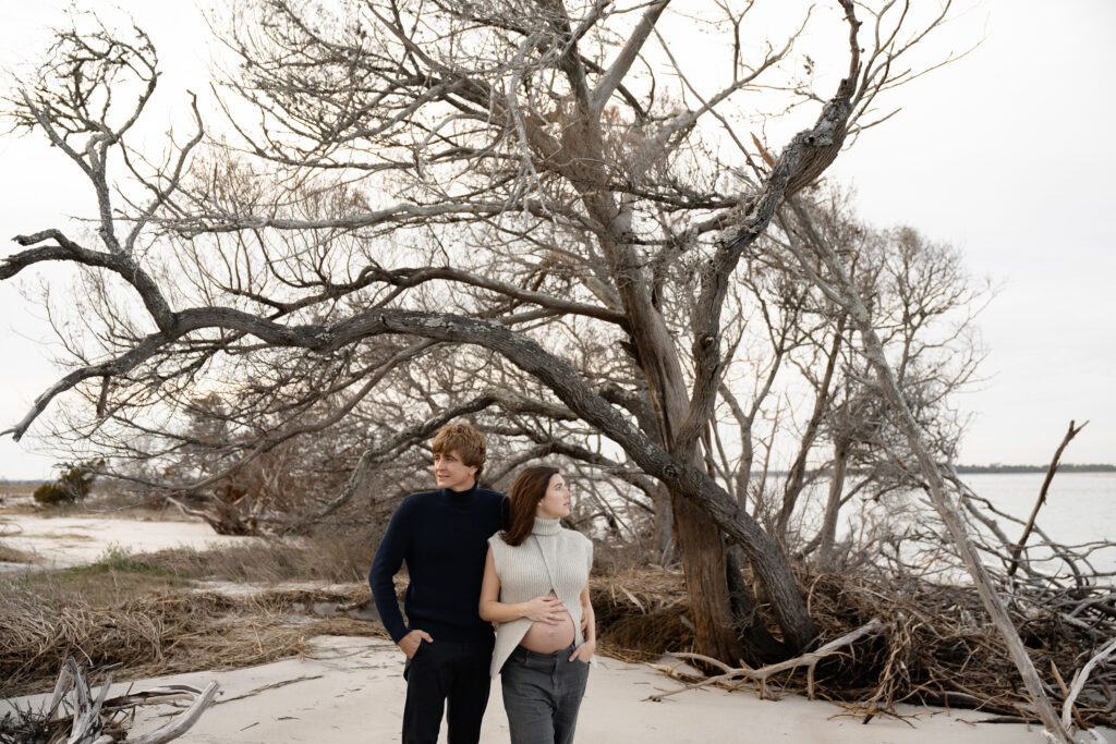 A couple stands on a deserted beach with dry, leafless trees. The man, wearing a black sweater, has his arm around the woman in a gray sweater, who touches her pregnant belly. They look into the distance, surrounded by sparse vegetation and a calm sea.