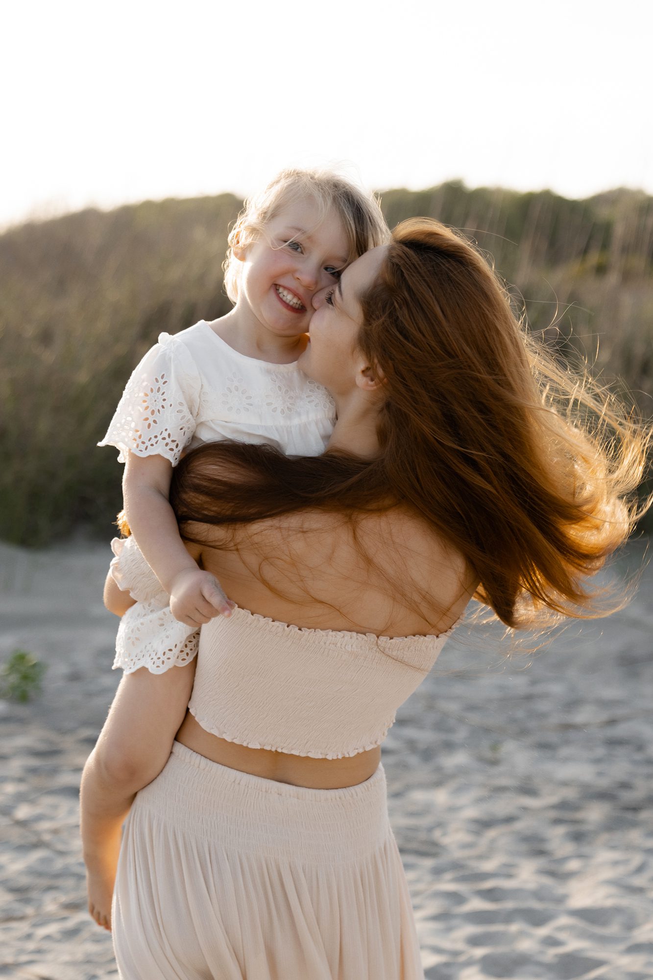 A woman with long brown hair is holding a smiling child with blonde hair wearing a white dress. They are both facing the camera, with the woman's face partially turned towards the child. Captured by a Charleston family photographer, they are outdoors on a sandy beach with vegetation in the background.