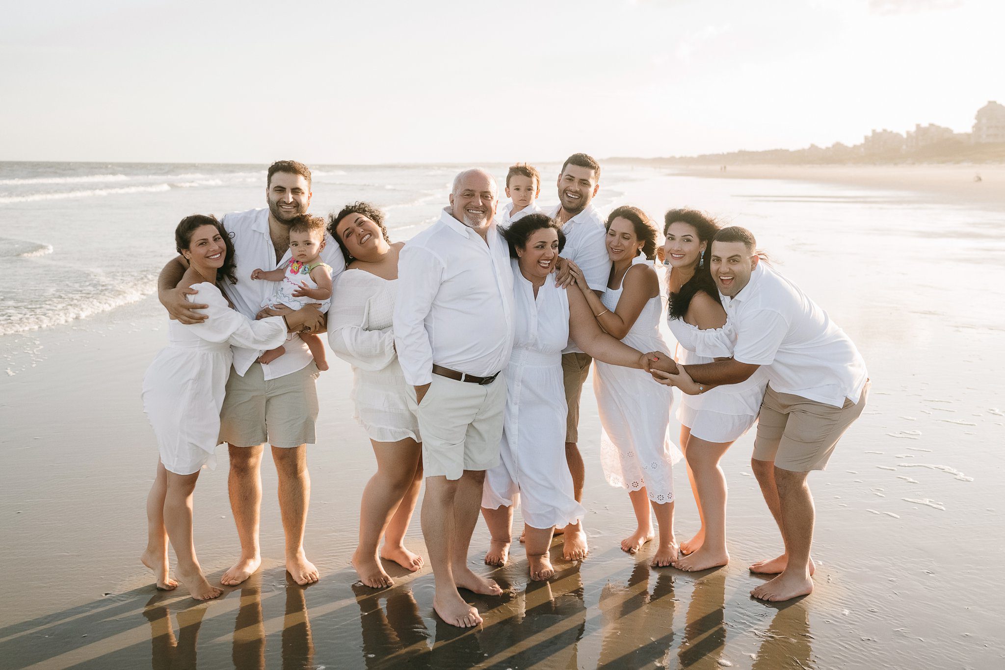 A group of eleven people, including three children, stand barefoot on a beach in light-colored clothes. The adults are gathered closely, smiling and embracing each other as the sun sets over the ocean in the background. Captured by a talented Kiawah Island photographer, the moment radiates warmth and connection.