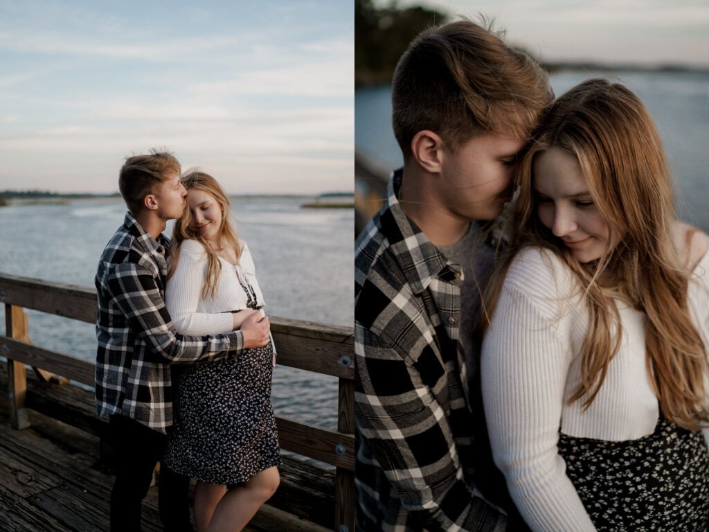 A couple stands on a wooden dock by the water at sunset during their maternity photoshoot in Charleston, SC. The man embraces the woman from behind, who cradles her pregnant belly. Both are smiling softly with closed eyes. The woman wears a white sweater and floral skirt, while the man wears a plaid shirt and jeans.