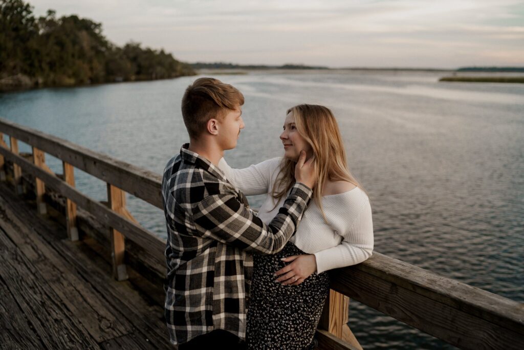 A young couple stands on a wooden boardwalk over a body of water, with trees in the background. The man, wearing a plaid shirt, gently touches the woman's face. The woman, wearing a white sweater and a patterned skirt, smiles at him lovingly.