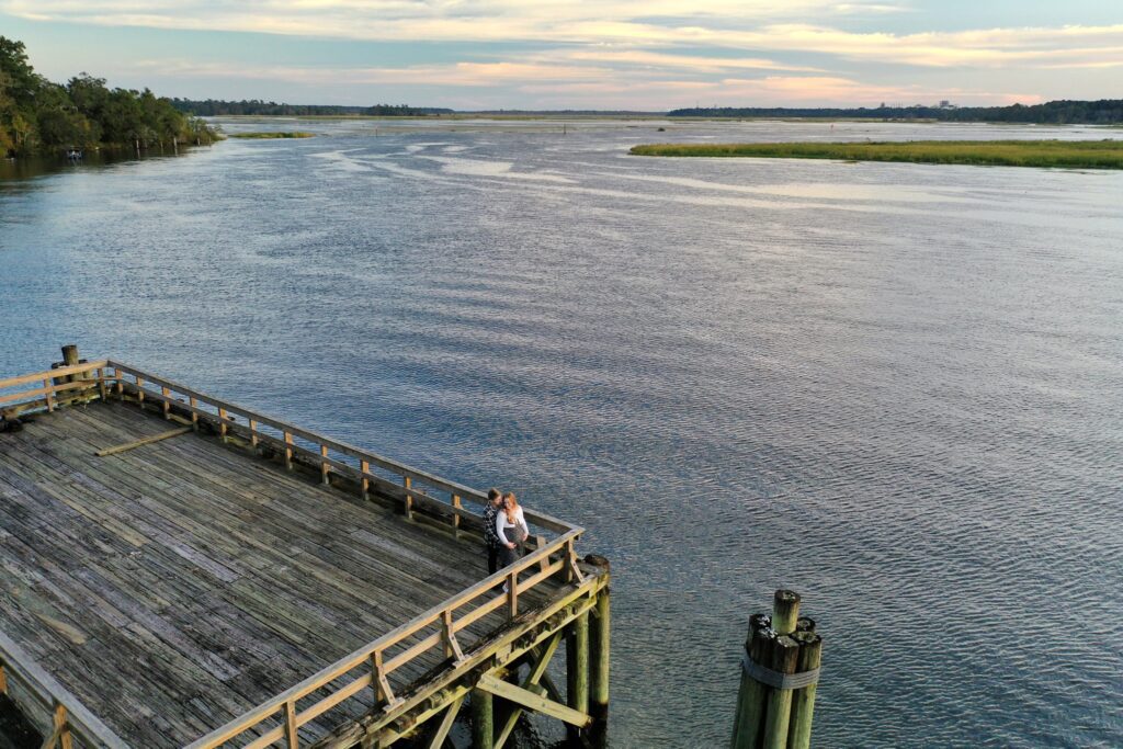 A couple stands at the edge of a large wooden dock, overlooking a serene, wide river. The sky is painted with light clouds and the horizon stretches with green vegetation. The atmosphere is calm and picturesque as they embrace during their maternity photoshoot in Charleston, SC.