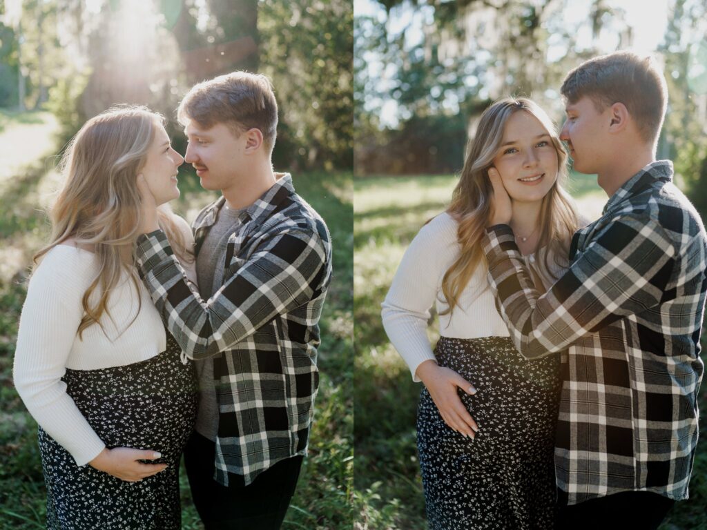 A couple stands in a grassy field with sunlight filtering through the trees. The man, in a plaid shirt and dark pants, holds her close. The woman, pregnant and wearing a long-sleeved top with a patterned skirt, cradles her belly. They gaze at each other lovingly during their maternity photoshoot in Charleston SC.