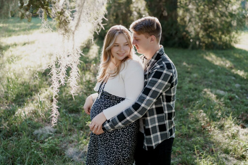 A man and woman stand in a grassy area with trees. The man, in a black and white flannel shirt, stands slightly behind and embraces the woman, who is pregnant and wearing a white long-sleeve top and a black-and-white patterned dress. In this serene maternity photoshoot in Charleston, SC, they both smile subtly.