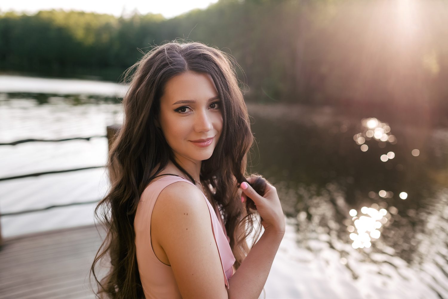 A woman with long, wavy hair stands on a dock by a serene lake, smiling gently at the camera. She is wearing a light-colored sleeveless top and holding some strands of her hair. The background features a sunlit forest and reflective water. Olga Chagarov, Charleston family photographer