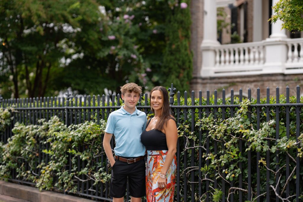 A teenage boy and a woman stand next to each other, smiling, in front of a wrought iron fence adorned with greenery during their Downtown Charleston Family Photoshoot. The boy is wearing a light blue polo shirt and black shorts, while the woman is dressed in a black top and a colorful skirt. A building with a white railing looms in the background.