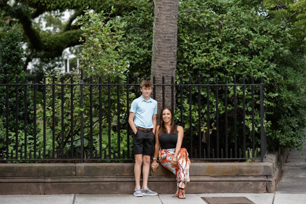 A young boy in a light blue polo shirt and black shorts stands next to a woman in a black top and colorful pants sitting on a ledge. They are outdoors on a sidewalk, framed by lush greenery behind a black metal fence—capturing the essence of a Downtown Charleston family photoshoot.