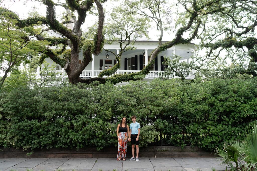 Two people stand on a sidewalk in front of a large, historic white house with tall, ivy-covered trees and lush greenery. The person on the left is wearing a black top and colorful pants; the person on the right sports a blue shirt and shorts—a perfect scene for Downtown Charleston family photoshoot.