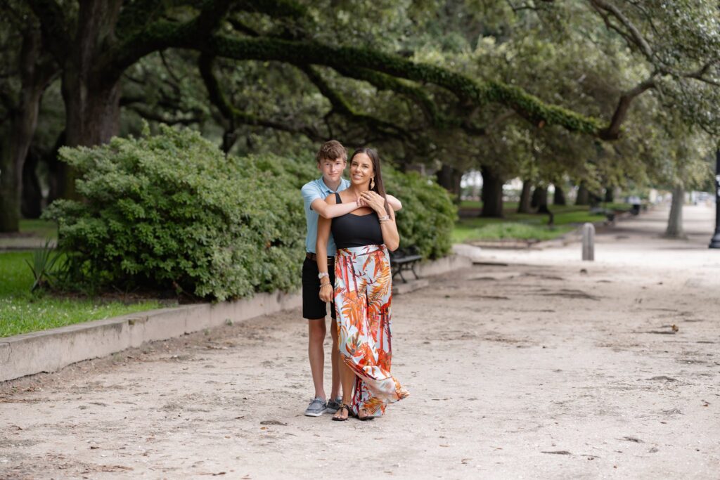 A woman in a colorful floral dress stands on a sandy path, smiling, with a young boy in a blue shirt embracing her from behind. They are surrounded by lush green trees and greenery, creating the perfect backdrop for their Downtown Charleston family photoshoot.