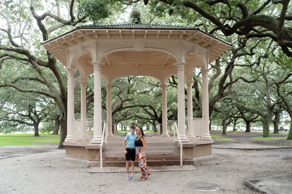 A couple stands in front of a large, cream-colored gazebo surrounded by lush green trees with sprawling branches in a park. The woman wears a long colorful dress, and the man is in a blue shirt and black shorts. The atmosphere is serene and picturesque.