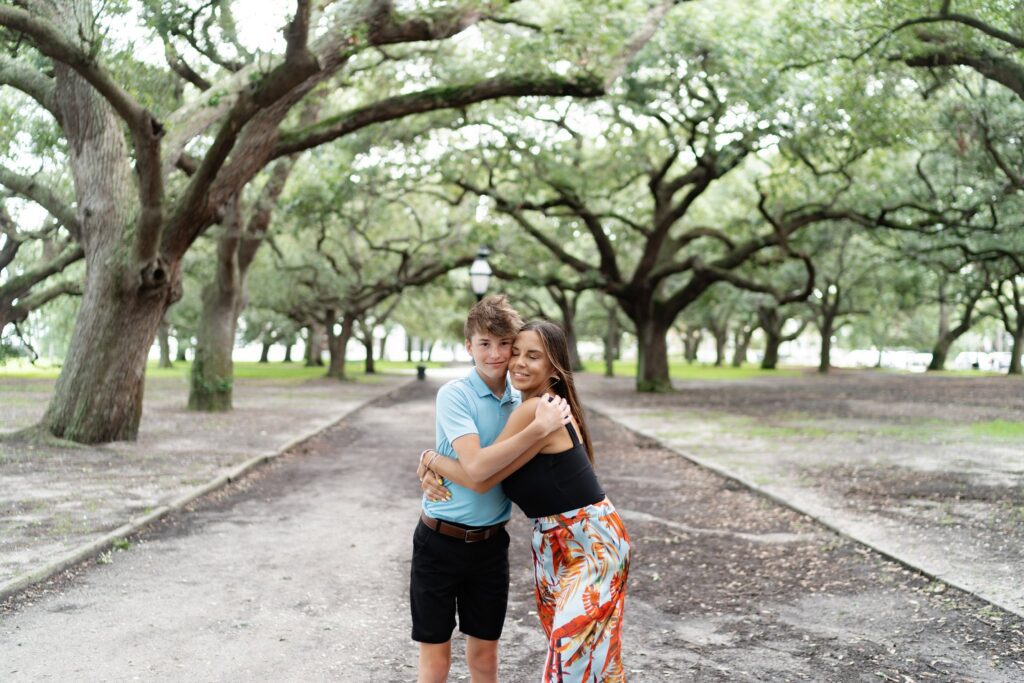 A young couple hugs on a tree-lined path in a park, reminiscent of a Downtown Charleston family photoshoot. The man wears a light blue shirt and black shorts, while the woman sports a black top and colorful floral pants. They smile and embrace under large trees curving overhead, creating a picturesque tunnel of greenery.