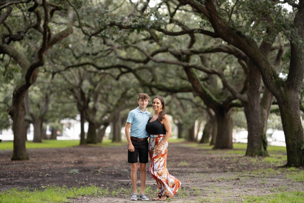 A young man and woman stand closely together with trees arching overhead, forming a natural canopy. The man is wearing a light blue polo shirt and black shorts, while the woman is dressed in a black top and a long, colorful skirt. They both smile at the camera during their Downtown Charleston family photoshoot.