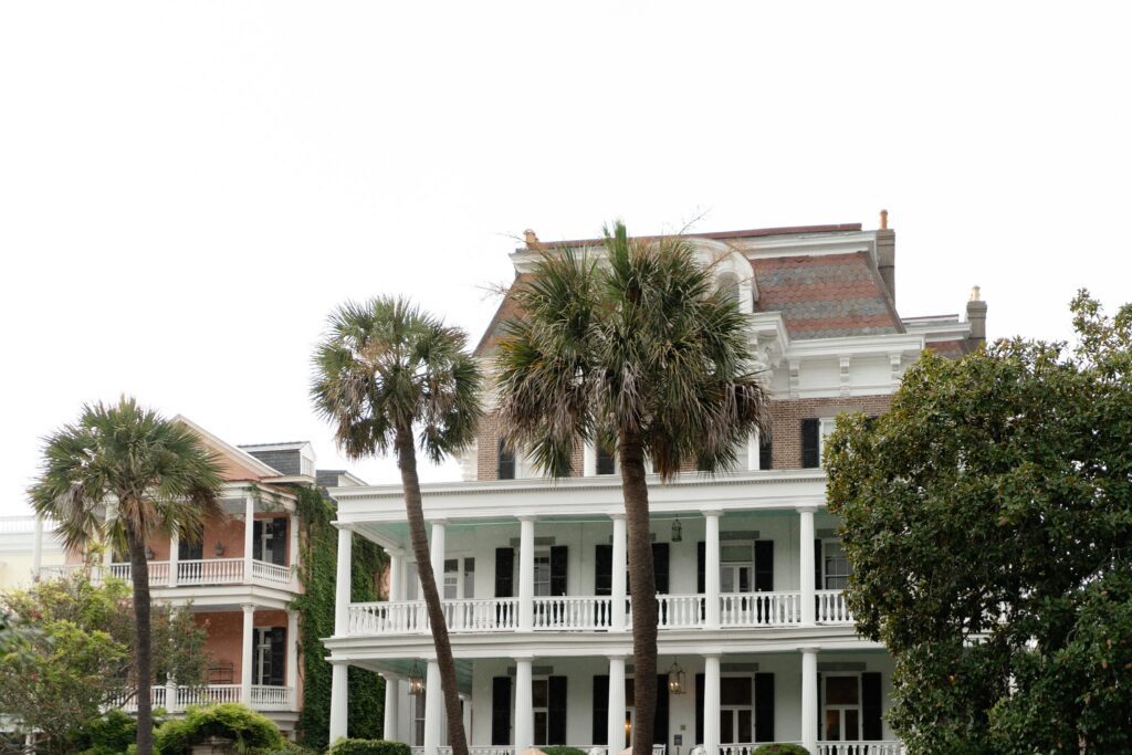 A large, multi-story house with white balconies and palm trees in front. The building has a mix of brick and white exterior, with lush greenery and trees surrounding it. The sky is overcast.