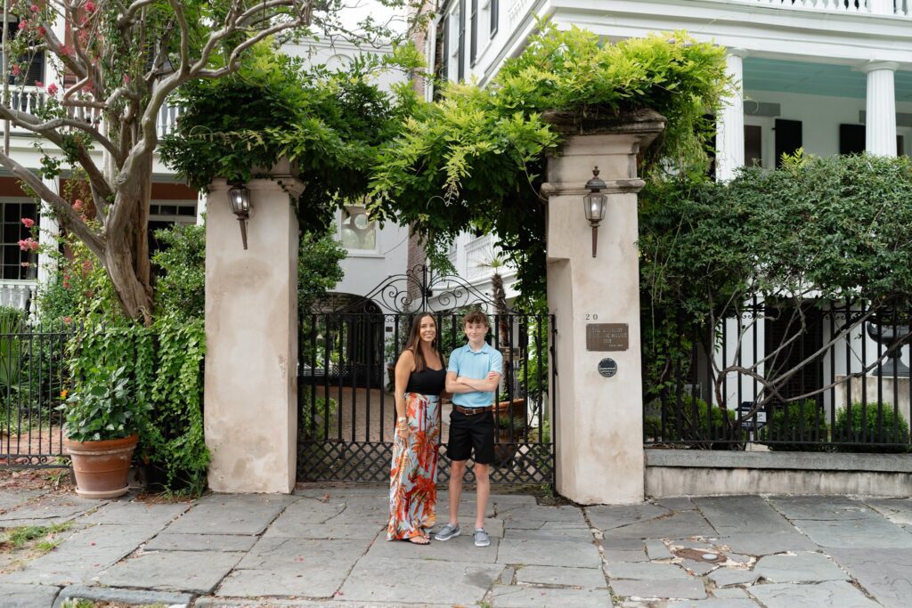 A woman and a boy stand in front of a gated entrance framed by tall stone pillars and greenery during their Downtown Charleston family photoshoot. The woman is wearing a black top and colorful skirt, while the boy is in a light blue shirt and black shorts. They pose before a large, white, columned house.