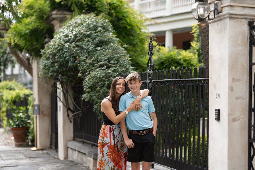 A woman and a boy stand in front of a black iron fence with a lush garden behind them, perfect for their Downtown Charleston family photoshoot. The woman wears a floral skirt and black top while the boy sports a light blue polo shirt and black shorts. Both are smiling, with the woman's arm around the boy's shoulders.