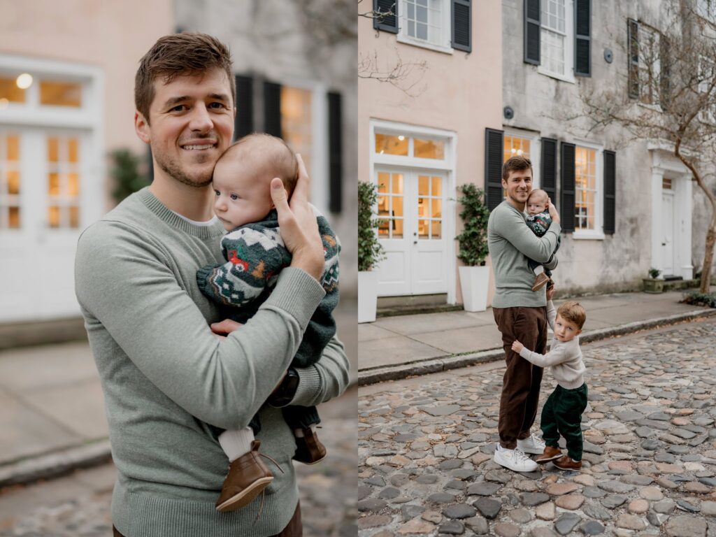 A man stands on a cobblestone street holding a baby, while a young child holds onto his leg. They are in front of a house with white doors and windows. The man is smiling, and both children are wearing sweaters.