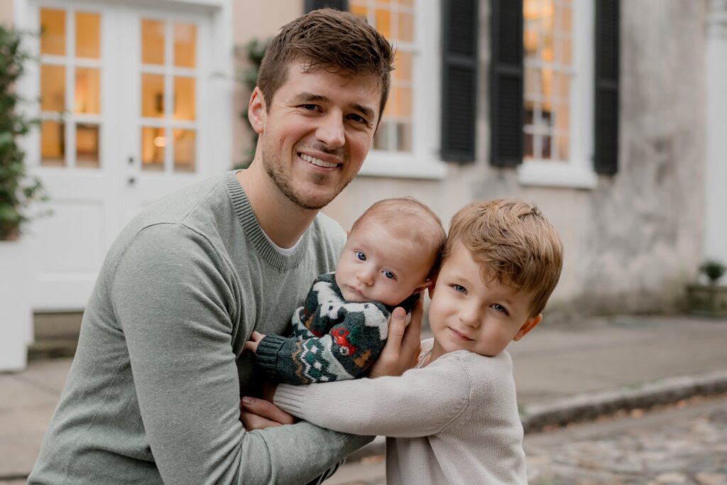 A man with short hair and a beard smiles while holding an infant in a patterned sweater. Next to him, a young child with short hair in a light sweater hugs the infant and smiles at the camera. They are outdoors in front of a building with white doors and windows, capturing the joy of Charleston mini sessions.