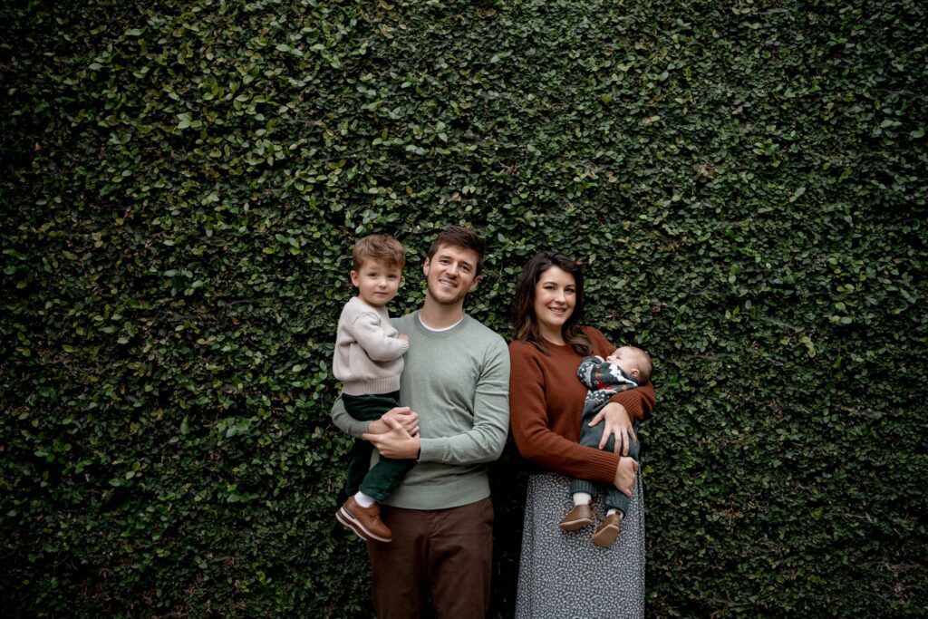 A family of four stands in front of a lush green ivy-covered wall during their Charleston mini session. The father holds a young boy, and the mother cradles a sleeping baby. Both parents are smiling and dressed in casual clothing while the young boy looks at the camera.