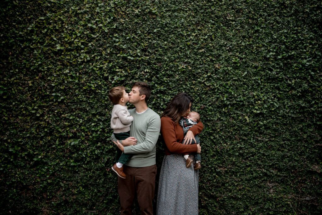 A man and woman stand in front of a lush green ivy wall. The man holds a small child who is kissing him on the cheek, while the woman, turned away from the man, cradles a baby in her arms. Both adults wear casual attire and the children are dressed warmly.