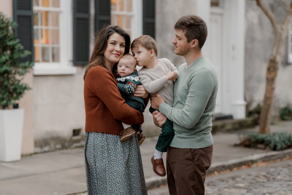 A family of four stands outside on a cobblestone street. The mother, wearing a brown sweater and patterned skirt, holds a baby. The father, in a light green sweater, holds their older child. Both parents look at each other while the older child looks at the baby—capturing this tender moment during their Charleston mini sessions.