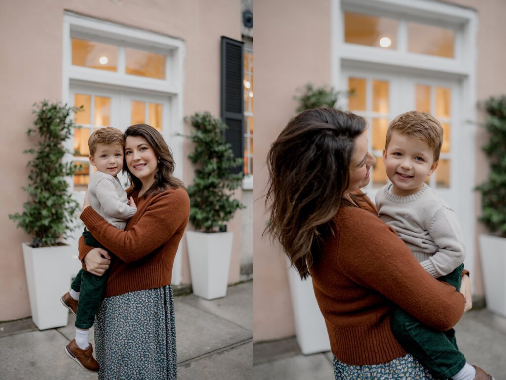 A woman wearing a rust-colored sweater and a blue patterned skirt holds a smiling young boy dressed in a gray sweater and green pants. They are standing outside in front of a light-colored building with large windows and potted plants, ready for their Charleston mini session. The background is out of focus.