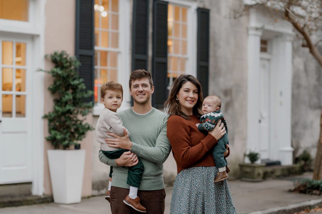 A family of four stands outside a building with large windows and plants. The father holds a young child wearing a white sweater, while the mother holds a baby in a patterned sweater. They both smile at the camera, dressed in casual attire, capturing the perfect moment during their Charleston mini sessions.