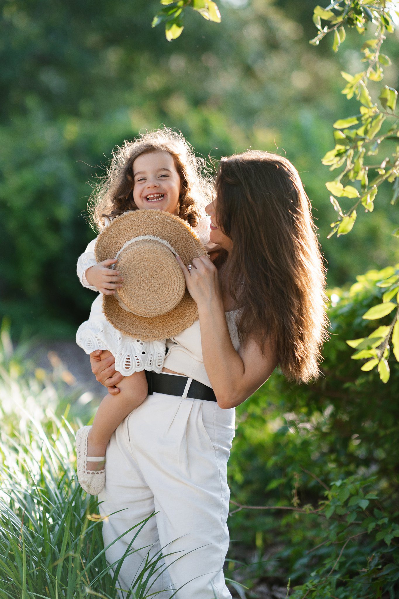 A woman holding a child in a sunlit garden. The child, with curly hair and a joyful smile, wears a white dress and holds a straw hat. The woman, in a sleeveless outfit with a black belt, embraces the child warmly. Captured by a Charleston photographer, both are surrounded by lush greenery.