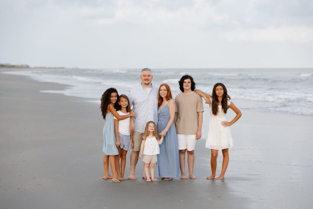 A family of seven poses on a sandy beach with gentle waves in the background. The group includes two adults and five children dressed in casual summer attire, with some barefoot. They stand closely together, smiling warmly at the camera. The sky is lightly overcast.