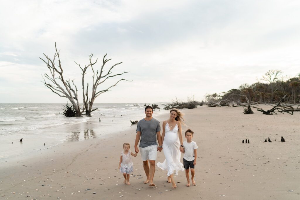 A family of four walks on a beach, holding hands. The parents are in the center with a little girl on the left and a boy on the right. The sandy shore is scattered with seashells and driftwood. In the background, leafless trees stand stark against a cloudy sky.