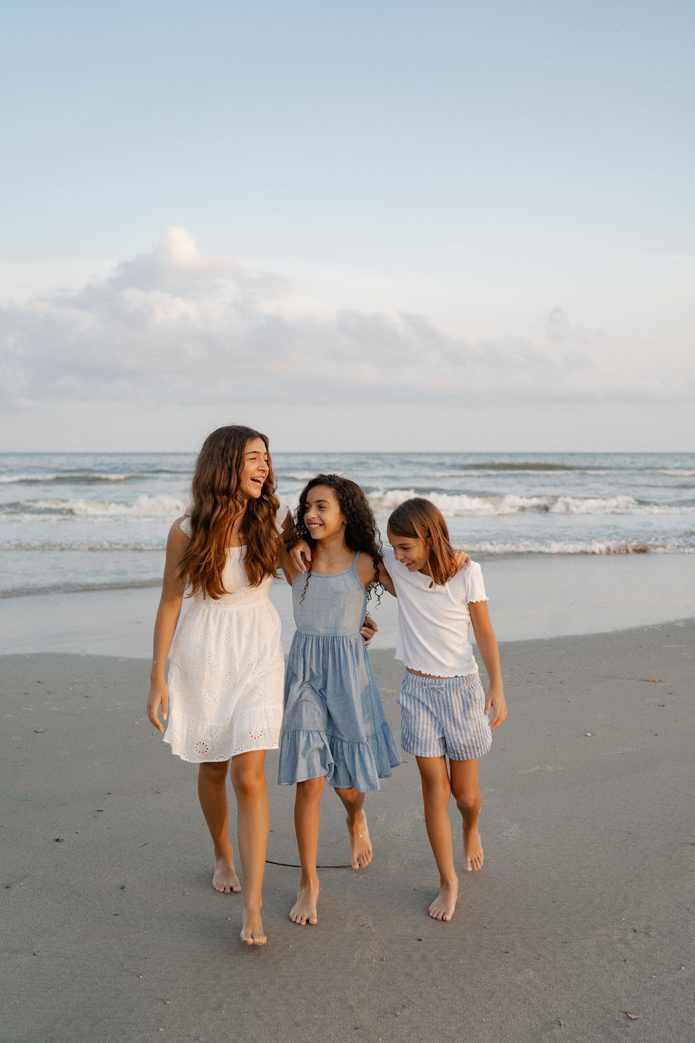 Three girls walk barefoot along a sandy beach at sunset, smiling and holding onto each other. The girl on the left wears a white dress, the middle girl wears a blue dress, and the girl on the right wears a white top with striped shorts. The ocean waves are in the background.