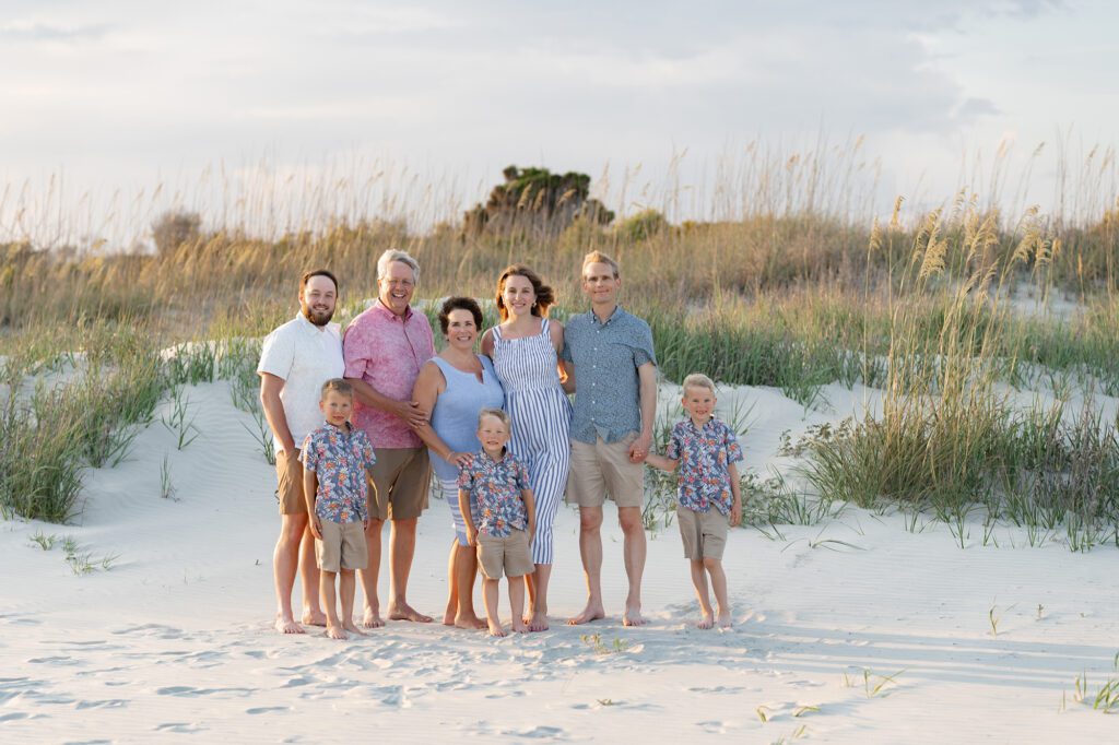 An extended family poses barefoot in the sand dunes at sunset on one of the best beaches for photos in Charleston, South Carolina. The group includes grandparents, parents, and young children dressed in coordinated summer outfits, surrounded by tall beach grass and soft white sand.