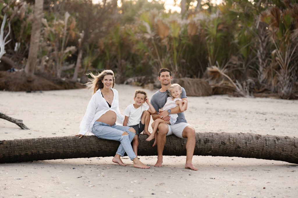 A pregnant woman, a man, and two young children sit together on a fallen palm tree on a sandy beach—one of the Best Beaches for Photos in Charleston SC—smiling at the camera with tropical trees in the background.