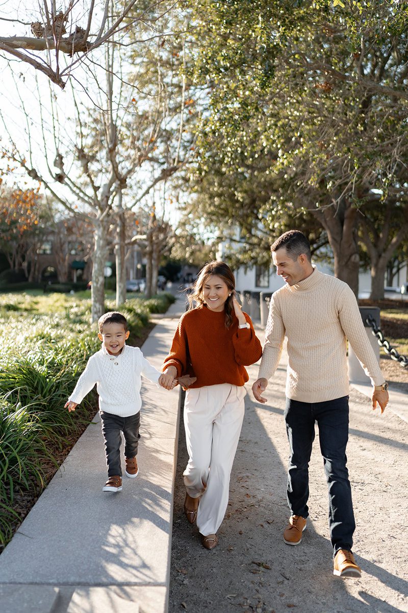 A smiling family of three—mother, father, and young son—walk outdoors on a sunny day. The parents hold hands with the boy, who is joyfully skipping. Trees and greenery line the path beside them.