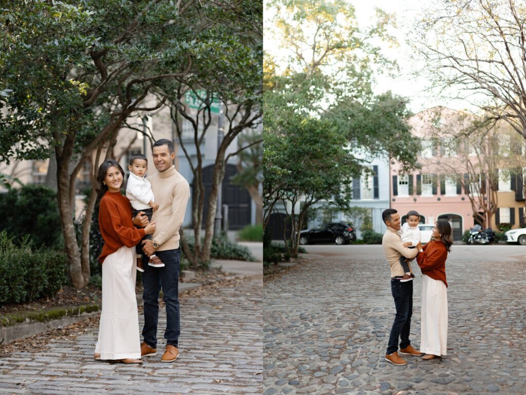 A young family of three stands on cobblestone streets in downtown Charleston, South Carolina, surrounded by trees and historic pastel homes. The parents smile warmly at the camera while holding their toddler, dressed in cozy neutral tones. Captured by photographer Charleston SC during a fall family photo session.