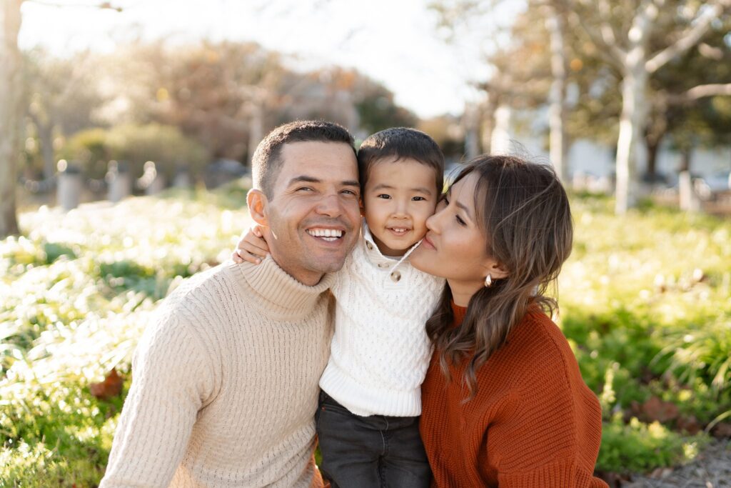 A smiling man, woman, and young boy sit close together outdoors. The woman kisses the boy on the cheek as he hugs both adults. Captured by a talented photographer in Charleston SC, sunlight filters through trees, creating a warm and happy scene.