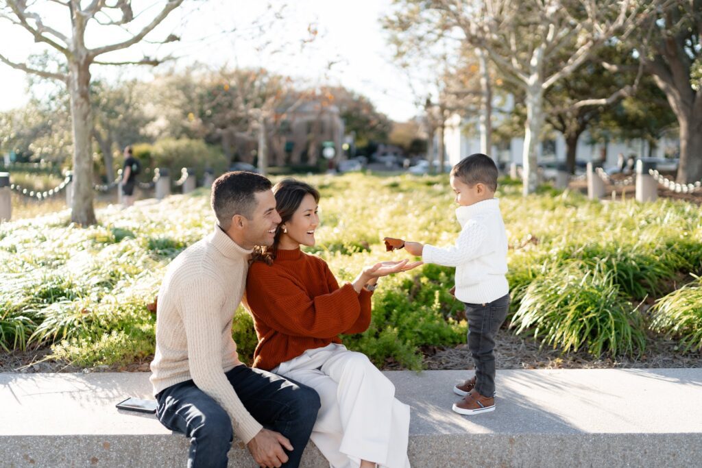 A young family sits on a stone bench in a park; the mother and father smile as their young son offers a brown leaf, all beautifully captured by a photographer Charleston SC. Lush trees and greenery complete the scene in the background.