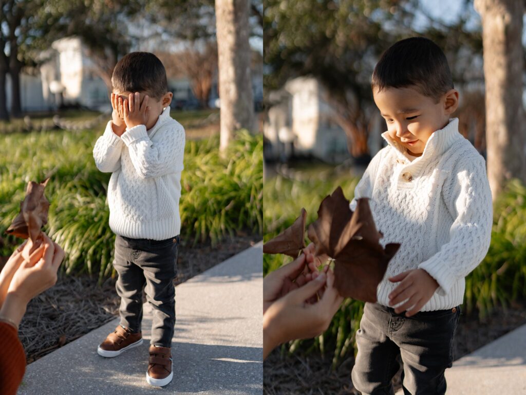 Split image of a young child outdoors: on the left, the child covers their face with their hands; on the right, they smile at a large brown leaf being handed to them by an adult. The child wears a white sweater and dark pants.