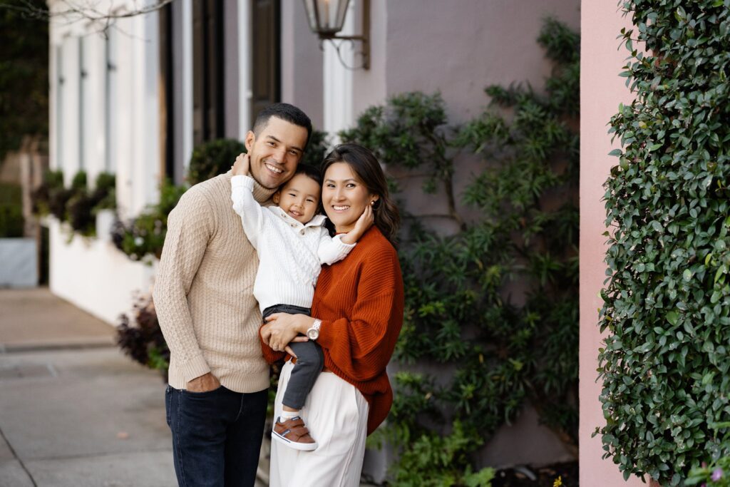 A smiling family of three stands outside by greenery. Captured by a photographer in Charleston, SC, the father and mother wear cozy sweaters as the mother holds their young child, all happily looking at the camera.