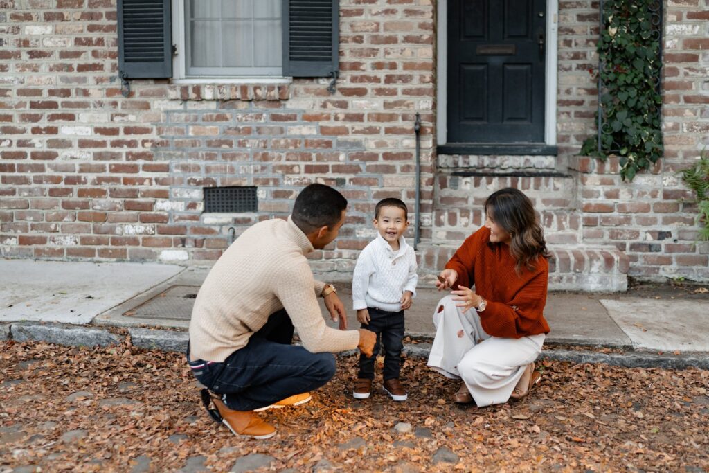 A toddler smiles brightly while standing between his parents in front of a historic brick home in downtown Charleston, SC. The parents kneel beside him, sharing a playful moment during their fall family session. Captured by photographer Charleston SC, this candid shot highlights the charm of Charleston’s architecture and the warmth of family connection.