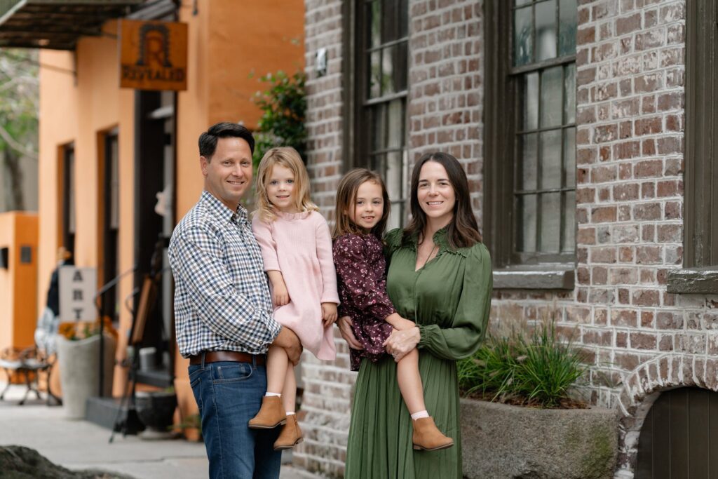 A family of four poses and smiles outside on a city sidewalk during mini sessions in Charleston, SC. The father wears a checked shirt and jeans, the mother is in a green dress, and their daughters sport pink and purple dresses with brown boots.