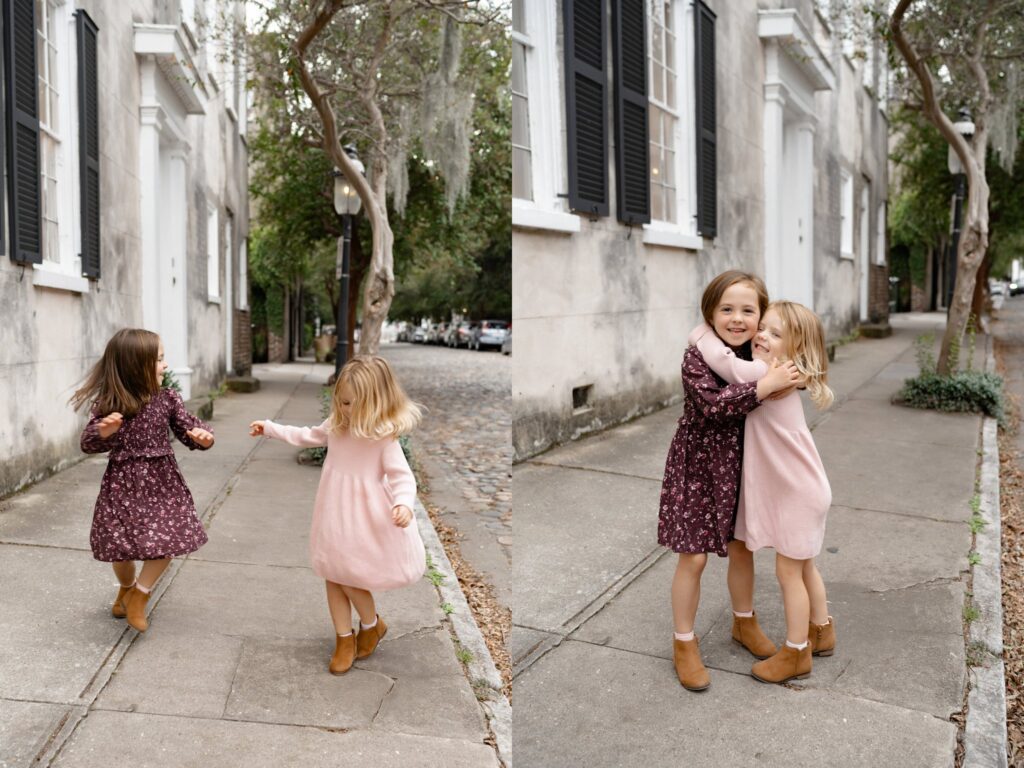 Two young girls, one in a floral dress and one in a pink dress, play and hug on a quiet, tree-lined sidewalk with historic buildings in the background.