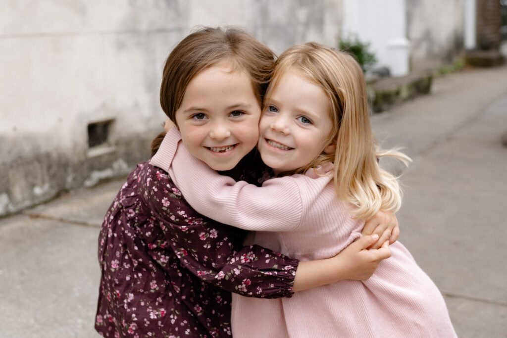 Two young girls smiling and hugging each other outdoors during mini sessions Charleston SC. One wears a dark floral dress, the other a light pink dress, as they stand on a sidewalk near a building wall.