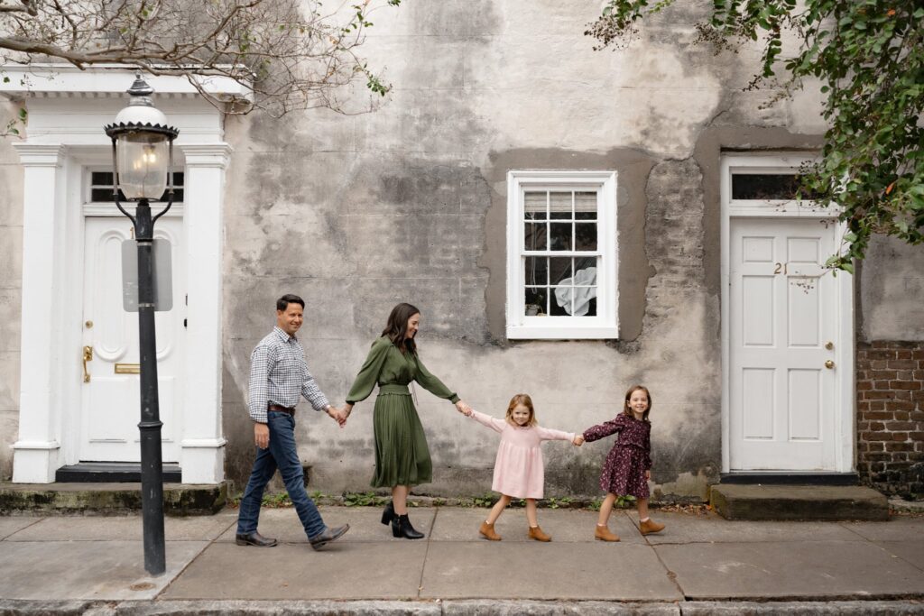 A family of four, holding hands and smiling, walks in front of an old textured building. The quiet, tree-lined street in Charleston SC is perfect for mini sessions capturing joyful moments and colorful outfits.