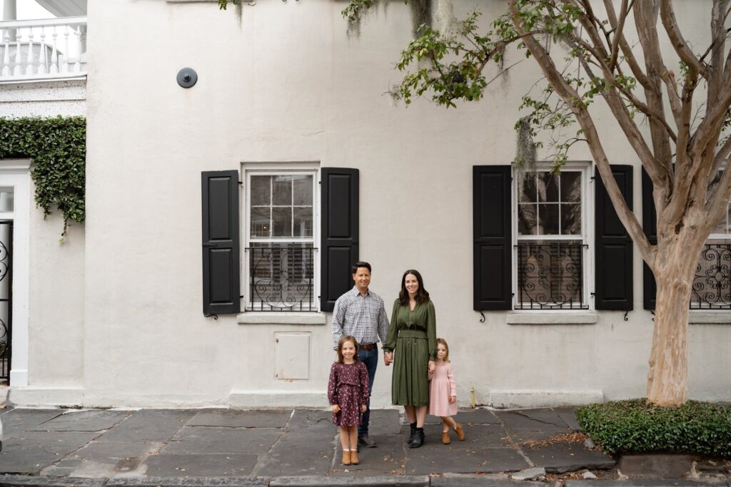 A family of four stands in front of a white building with black shutters. The parents hold hands, with two young daughters in patterned dresses beside them. A tree is on the right, and the sidewalk is in the foreground.