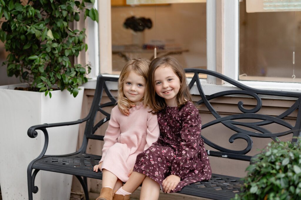 Two young girls sit closely together on a black decorative bench outdoors during mini sessions Charleston SC. One wears a pink dress, the other a dark purple floral dress. They're smiling happily, with plants and a window in the background.