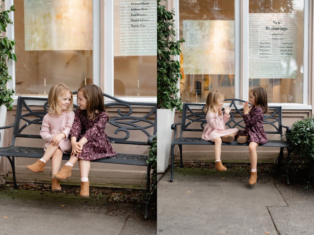 Two young girls sit together on a black metal bench outside a building. One wears a pink dress and tan boots, the other wears a dark purple floral dress. They are smiling and talking, surrounded by greenery.