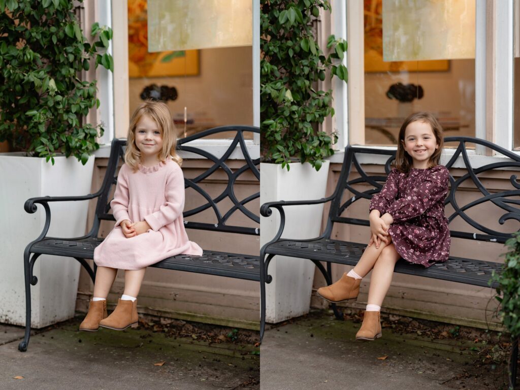 Two young girls sit on a black metal bench outside a building during mini sessions Charleston SC. The girl on the left wears a pink dress and brown boots, while the girl on the right sports a maroon floral dress and brown boots. Both are smiling.