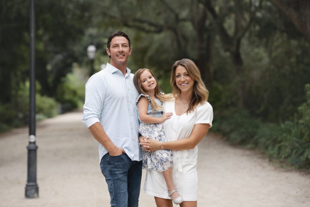 A man, woman, and young girl pose together outdoors on a tree-lined path, captured by a family photographer in Charleston SC. The man wears a light blue shirt and jeans, the woman is in a white dress, and the girl sports a floral dress as they all smile.
